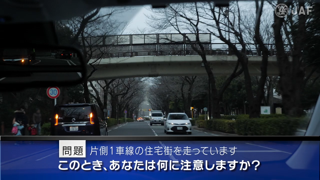 実写版危険予知「SCENE53　住宅街編 その6 ～片側１車線の住宅街を走っています。左側には集合住宅があり、右側には大きな公園があります。対向車が来ていますが道幅に余裕があるので、左側に止まっている車の側方を通過しようと思います。～」